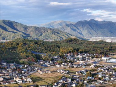 Bir kasabanın arka planında bir dağ sırası görülebilir. Kasaba evler ve binalarla dolu ve gökyüzü bulutlu.