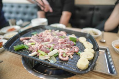 Izgarada bir tabak et pişiyor. Et küçük parçalara ayrılır ve sebzelerle çevrilidir. Sahne bir restoranda geçiyor, bir grup insan bir masanın etrafında oturuyor.