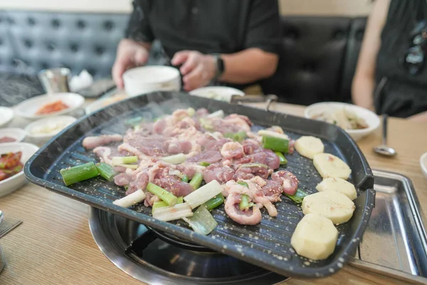 Izgarada bir tabak yemek pişiriliyor. Yiyecek, soğan ve patates de dahil olmak üzere et ve sebzelerin bir karışımıdır. Sahne bir restoranda geçiyor, birkaç kişi yakınlardaki bir masada oturuyor.