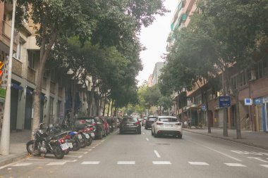 Yan tarafında arabalar ve motosikletler olan işlek bir cadde. Arabaların çoğu beyaz ve motosikletlerin çoğu siyah. Sokaklar ağaçlar ve binalarla dolu.