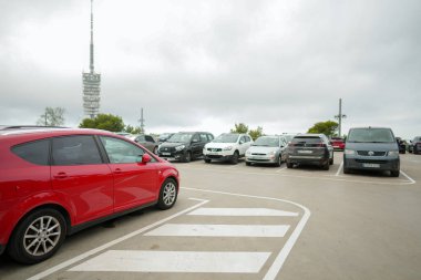 A parking lot with a red car in the foreground and a white van in the background. The parking lot is full of cars and the sky is cloudy
