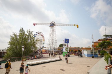 Bir grup insan, arka planda dönme dolap olan bir parkta dolaşıyorlar. Gökyüzü bulutlu ve atmosfer canlı.