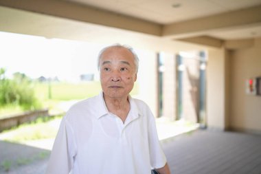 Beyaz gömlekli ve gri saçlı yaşlı bir adam bir binanın önünde duruyor. Üzgün ve yalnız görünüyor.