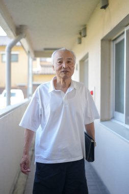 Beyaz gömlekli ve siyah şortlu bir adam koridorda yürüyor. Siyah bir çanta ve evrak çantası taşıyor.