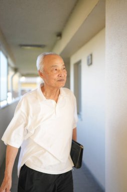 Beyaz gömlek ve siyah şort giyen yaşlı bir adam koridorda yürüyor. O, siyah bir çanta ve bir kitap taşıyor. Adamın acelesi var, muhtemelen bir toplantıya ya da randevuya gidiyor.