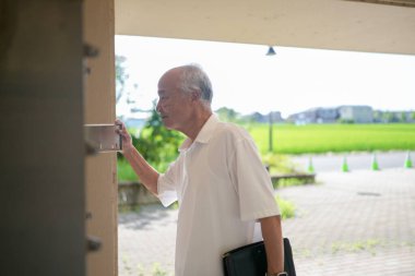 Beyaz gömlekli bir adam binanın dışında duruyor ve bir kapıya bakıyor. Elinde bir evrak çantası var ve acelesi var.