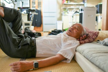 Yaşlı bir adam yastıkla yatakta yatıyor. Beyaz gömlek ve gri pantolon giyiyor.