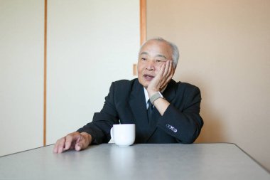 Takım elbiseli bir adam önünde bir fincanla masada oturuyor. Yorgun görünüyor ve yüzünü ovuyor.