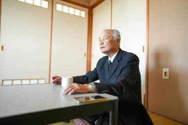 Takım elbiseli bir adam bir fincan kahveyle masada oturuyor. Telefonuna bakıyor.