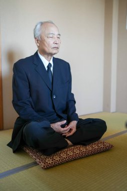 Takım elbiseli bir adam bir yastığın üzerinde oturuyor ve elleri önünde sıkıca bağlı. Derin düşüncelere dalmış ya da meditasyon yapıyor.