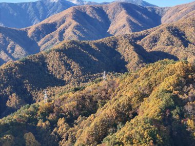 Dağlar ağaçlarla kaplı ve yapraklar renk değiştiriyor. Gökyüzü açık ve güneş parlıyor.