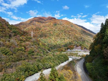 İçinden yol geçen bir dağ sırası. Yol ağaçlarla kaplı ve yapraklar renk değiştiriyor. Gökyüzü açık ve güneş parlıyor.