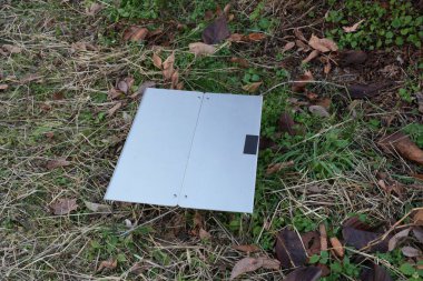 Çimenli bir alanda yerde gümüş bir kutu duruyor. Kutu açık ve üzerinde siyah bir etiket var. Sahne huzurlu ve sakin, önplandaki tek nesne kutu.