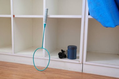 Bir tenis raketi bir odada bir rafa yaslanıyor. Raket yeşil ve mavi saplı. Duvarda mavi bir çanta asılı.