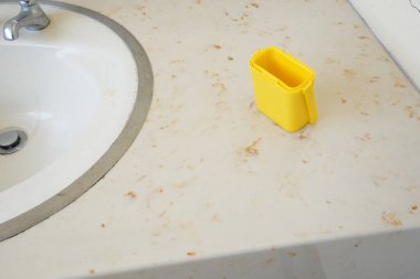 Sarı bir kap kirli bir tezgahın üzerinde lavabonun yanında duruyor. Lavabo kirli ve etrafında sarı bir halka var.