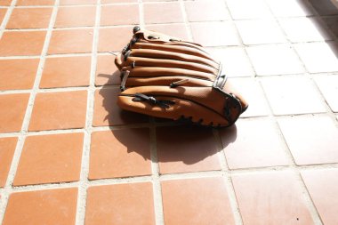 Bir beyzbol eldiveni fayanslı zeminde yatıyor. Eldiven kahverengi ve siyah bir kayışı var. Eldiven kullanılmıyor ve o zarar görmüş.