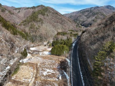 Karlı bir dağ vadisinde bir yol akıyor. Yol boş ve ağaçlar çıplak. Gökyüzü açık ve güneş parlıyor.