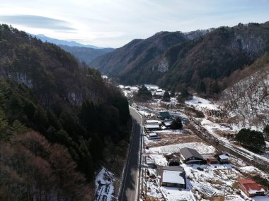 Karlı bir dağ sırasının içinden geçen bir yol. Yol boş ve evler küçük.