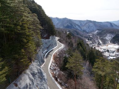 Arkasında ağaçlar olan karlı bir dağ yolu. Yol dolambaçlı ve dar, ağaçlar uzun ve yeşil. Sahnenin huzurlu ve huzurlu bir havası var.