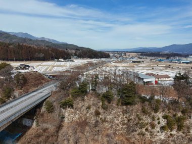 Karlı bir tarlanın arka planında bir dağ sırası görülebilir. Gökyüzü açık ve mavi, ve karla kaplı zemin ağaçlarla dolu.