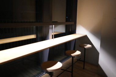 Tahta tezgahı ve üç taburesi olan küçük, loş bir oda. Oda boş ve ışık açık.