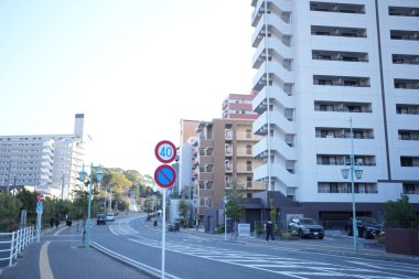 Günışığı şehir caddesi ve modern apartmanlar. Açık gökyüzü ve sakin trafik akışı yerleşim alanlarının önünden geçiyor. Yayalar yaya geçitlerinin yanında yürür, telaşsız ve düzenlidir.