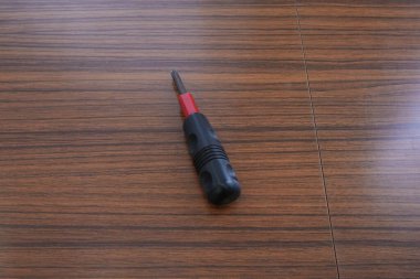 Alet parlak ahşap yüzeyde duruyor. Siyah tutuş, kırmızı uç, basit tasarım. Temiz bir arkaplan nesnelerin berraklığını vurgular. DIY, onarım veya bakım temaları için ideal