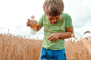 Yeşil tişörtlü, kirli bir çocuk buğday tarlasında dikilirken üzerine içki döker. Kırsal kesimde. Mutluluk ve eğlence konsepti.