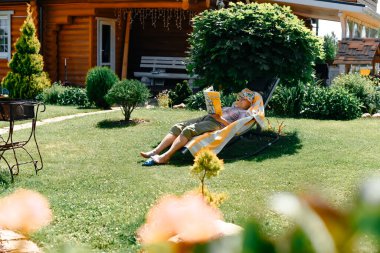 Grandmother in the summer at the dacha lies on a sunbed in a poneme reading a book. Resting lying near the trees. Fresh air and relaxation concept. clipart