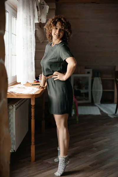 A young attractive girl in a dark green dress with curly hair stands in a room by the window. Sunlight. Curly short hair. Beauty and fashion concept. Wooden house.