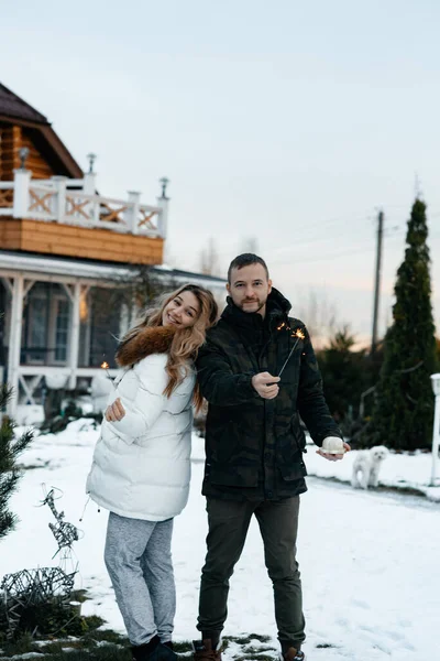 Husband and wife stand in the winter near the fir holding the Bingala fires. New Year\'s Holidays. The concept of happiness and love. Snowy country house. It\'s snowy time.