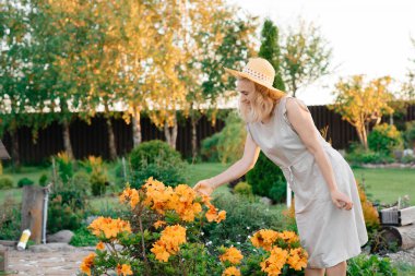 Beyaz elbiseli ve hasır şapkalı tatlı bir kız güneşli bir havada çiçek açan sarı çiçeklerin yanında yürüyor. Peyzaj.