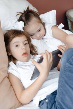 Küçük bir oğlan ve özel ihtiyaçları olan bir kız tabletle oynayan bir odada yatıyorlar. Tabletli beyaz gömlekli bir çocuk. Özel ihtiyaçları olan çocuklara bakma kavramı. Uzayı kopyala Tonlama.