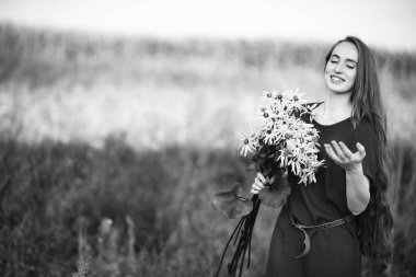 Tarlada bir buket çiçek toplamış uzun saçlı güzel bir kızın siyah beyaz fotoğrafı. Kırsalda yürü. Bir buket papatyası olan bir kız..