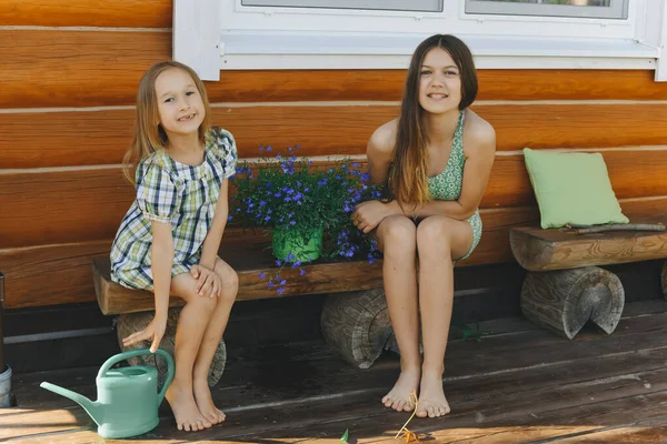Две Маленькие Девочки Летних Платьях Сидят Скамейке Рядом Деревянным Домом — стоковое фото