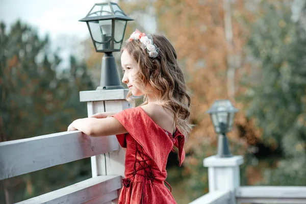 オープンバルコニーに沿って歩く彼女の頭の上にフープを持つ長い赤いドレスの美しい少女 王女様 黄色の紅葉 — ストック写真