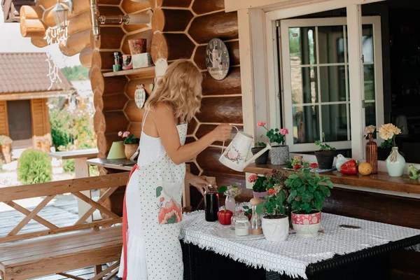 白いドレスを着た金髪の女の子がテラスの鍋に花を散水しています ホームプラントケアの概念 テラスの花 — ストック写真