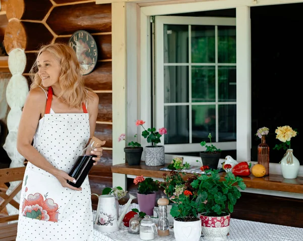 A girl in a white dress and an apron stands on the terrace near flowers holding a bottle in her hands. flowers on the terrace.  Home plant care concept.