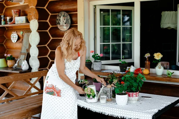 白いドレスを着た女の子とエプロンは テラスや窓辺のポットにある家の花を世話します ホームプラントケアの概念 テラスの花 — ストック写真