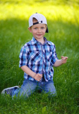 Küçük bir çocuk mavi ekose gömlekli parkta yeşil çimenlerin üzerinde oturuyor