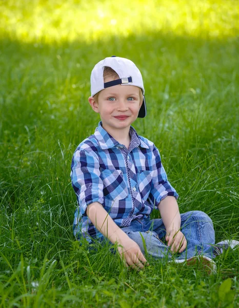 Un petit garçon en chemise à carreaux bleue est assis sur l'herbe verte dans le parc — Photo