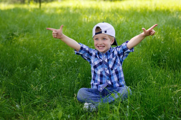 A little boy in a blue plaid shirt is sitting on the green grass in the park — Stock Photo, Image