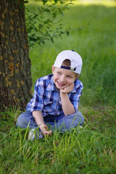 A little boy in a blue plaid shirt is sitting on the green grass in the park — Stock Photo, Image