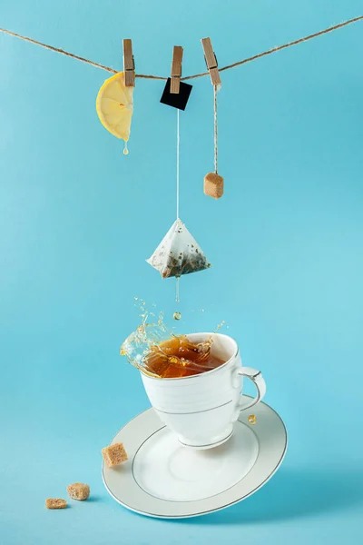 Torebka herbaty, cytryna i cukier wiszące na linie nad rozpryskiwaniem herbaty na niebieskim tle. Kreatywna koncepcja martwej natury. — Zdjęcie stockowe