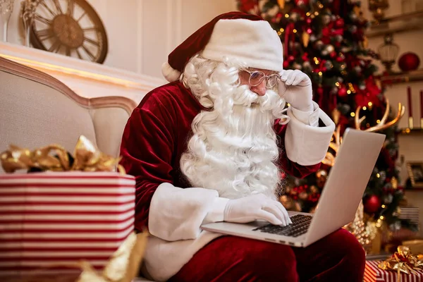 Santa Claus sitting at his home and reading email on laptop with hristmas requesting or wish list near the fireplace and tree with gifts. New year and Merry Christmas ,happy holidays concept