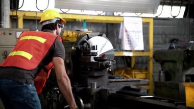 Güvenlik Üniforması Giyen, Gözlüklü ve Sert Şapkalı İşçi çelik makinesini kontrol ediyor. Arka planda Odaklanmamış Büyük Endüstriyel Fabrika.