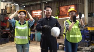 Asyalı fabrika işçisi ve kadın işçilerden oluşan bir grup kask emniyetini takıyor ve güvenle ayakta duruyor ve gülümsüyor. Sanayi fabrikasının arka planında.