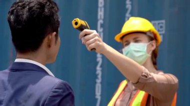 Maskeli bir fabrika işçisi ve bir virüs salgını sırasında vücut ısısını ölçmek için iş yerlerinde termometre kullanıyor. Ve işçi fabrikanın içinden geçiyor..