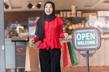 Kırmızı elbiseli, mutlu yüzlü Müslüman kadın alışveriş çantası taşıyor ve dükkândan açık tabelayla çıkıyor. Alışveriş festivali mevsiminde ve yeni yılda insan etkinliği kavramı.
