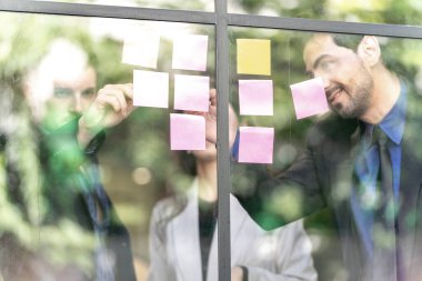İş adamları ofisteki cam pencere duvarına yapışkan notlar yapıştırarak beyin fırtınası yapıyorlar. Pencere camı gün ışığını ve çevreyi yansıtıyor. Yeni fikir için yaratıcı grup girişimi iş toplantısı.